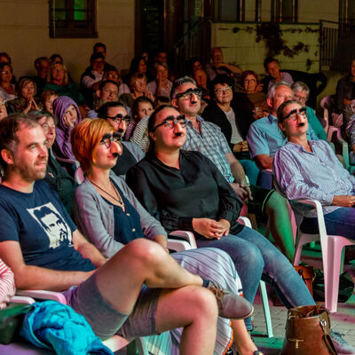 In mehreren Reihen sitzende Menschen, die aufmerksam der Band zuhren. Vier Personen in der ersten Reihe tragen Faschingsbrillen mit falschen Nasen.