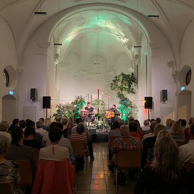 Musikkonzert in einer Kapelle. Die Besucher blicken auf die Musiker Susi Raith und Mathias Kellner, die auf einer bunt beleuchteten und dekorierten Bhne Gitarre spielen.