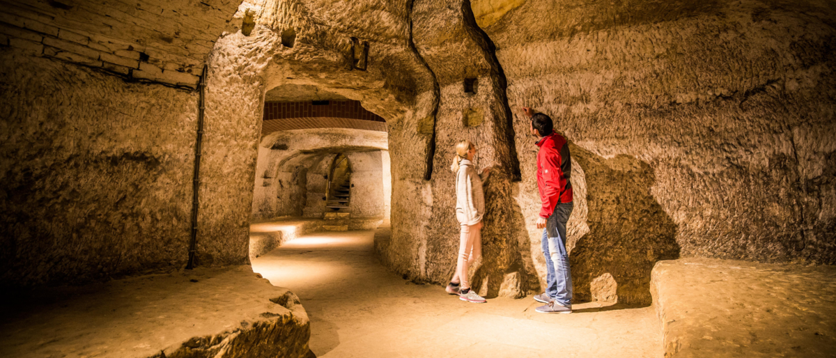 Fotot des Felsenkellertunnels. Ein Mann und eine Frau stehen an der braunen-orangenen Wand und betrachten das Gestein. Der Tunnel ist hell erleuchtet.