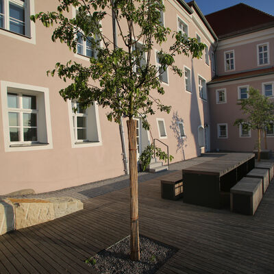 Bild vergrößern: Außenanlage der Volkshochschule.
Man sieht einen Holzboden mit einer länglichen Banklandschaft und einem Baum.