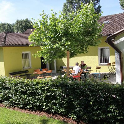 Bild vergrößern: Lesegarten in der Stadtbibliothek. Es stehen viele Tische und Stühle herum und in der Mitte zeigt sich ein Baum in voller Pracht.