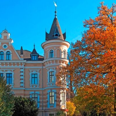 Bild vergr&ouml;&szlig;ern: Das Geb�ude des Oberpf�lzer-K�nstlerhauses. An der rechten Seite t�rmt sich ein gro�er Baum mit roten Bl�ttern. Im Hintergrund das Haus mit vielen Fenstern und einem Turm.