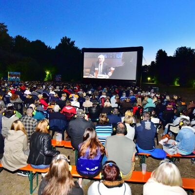 Bild vergrößern: Foto des Open-Air Kinos im Stadtpark bei Nacht. Man sieht über die Köpfe des Publikums die Leinwand, auf welcher gerade ein Film läuft.