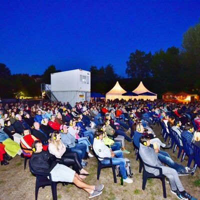 Bild vergrößern: Foto des Publikums beim Open-Air Kinos im Stadtpark bei Nacht. Im Hintergrund sieht man beleuchtete Zelte.