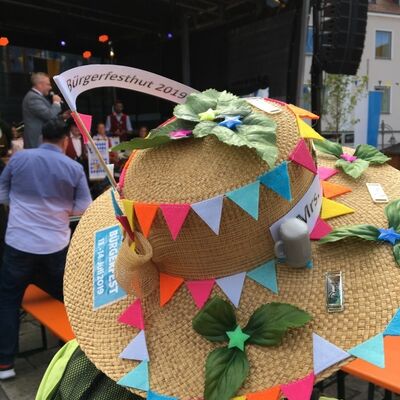 Bild vergrößern: Nahaufnahme eines mit bunten Fähnchen geschmückten Hutes von  Pallicura e.V.  Auf einem Fähnchen steht Bürgerfest 2019.