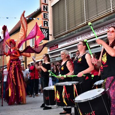 Bild vergrößern: Foto der Trommelgruppe Jalapenos und ein verkleideter Mensch des Rottaler Staatszirkus, die auf der Straße performen.