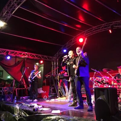 Bild vergrößern: Foto einer Band, die auf der Bühne am Kirchenplatz performt. Die Hintergrundbeleuchtung ist rot.