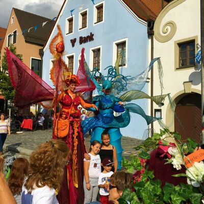 Bild vergrößern: Foto zweier großer Figuren, eine blau, eine rot, vor einer Gruppe von Kindern.