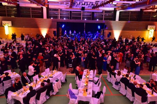 Bild vergrößern: Foto der gefüllten Tanzfläche in der Oberpfalzhalle. Im Vordergrund stehen drei Tafeln, die in weiß gedeckt sind. Die Menschen tanzen klassischen Tanz in festlicher Kleidung.