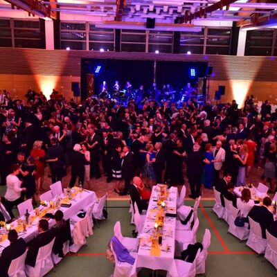 Foto der gefüllten Tanzfläche in der Oberpfalzhalle. Im Vordergrund stehen drei Tafeln, die in weiß gedeckt sind. Die Menschen tanzen klassischen Tanz in festlicher Kleidung.