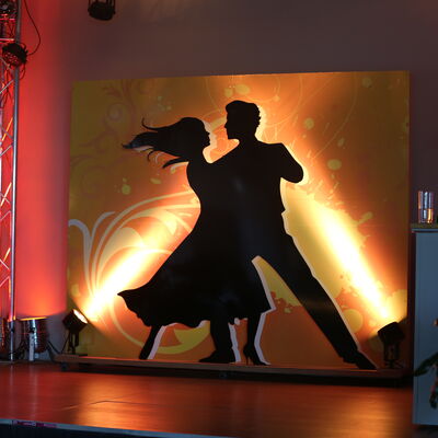 Foto vom Logo des Stadtballs. Logo: Eine Frau und ein Mann tanzen. Die Figuren sind komplett in schwarz auf einem gelben Hintergrund. Es wird angeleuchtet.