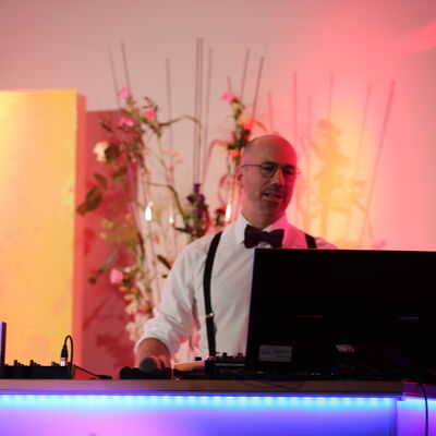 Foto des DJs Stefan Theuerl, der hinter dem DJ-Pult steht. Im Hintergrund ist ein großes Blumengesteck.