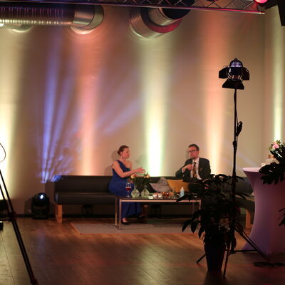 Die Moderatoren Anna Theuerl und Johannes Lohrer sitzen auf einem Sofa. Der Raum ist mit Licht dekoriert.