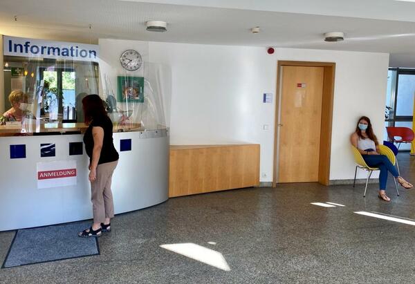 Bild vergrößern: Eine Person meldet sich bei der Information im Rathaus an. Eine weitere Person sitzt im Wartebereich bis ein Mitarbeiter der Stadtverwaltung in abholt.