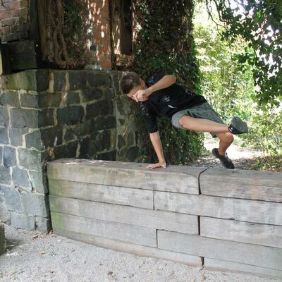 Ein Junge der sich mit einer Hand abstützt und über eine Mauer springt.
Überall ist es grün und es ist sonnig