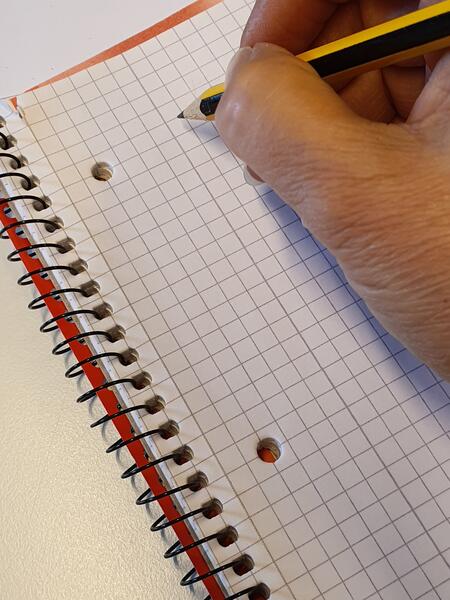Bild vergrößern: Kleiner Spiralblock mit Karomuster, Teil einer Hand mit Bleistift.