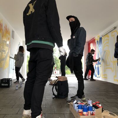Bild vergrößern: Zwei Graffiti-Profis, die den Workshop geleitet haben, stehen mit Mundschutz und Kapuzenpulli vor dem Kunstwerk.