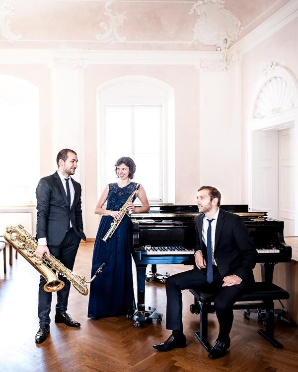 Bild vergrößern: Ein Gruppenfoto der Band Trio Etoiles. Sie tragen Abendgarderobe und stehen neben einem Piano.