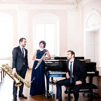 Ein Gruppenfoto der Band Trio Etoiles. Sie tragen Abendgarderobe und stehen neben einem Piano.