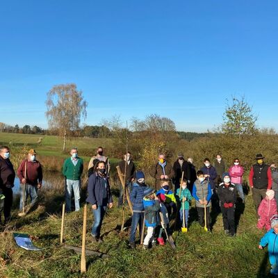 Bild vergrößern: Mehrere Erwachsene und Kinder die auf einer Wiese beim Bäume pflanzen helfen.