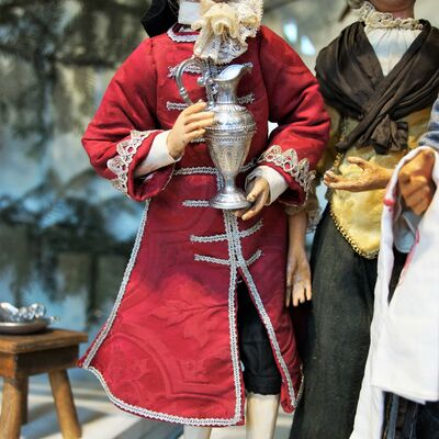 Bild vergrößern: Eine Figur aus der Krippe von der Familie Pöllmann,
es zeigt einen Mann in einem roten Gewand der einen silbernen Kelch in den Händen (vor der Brust) hält.