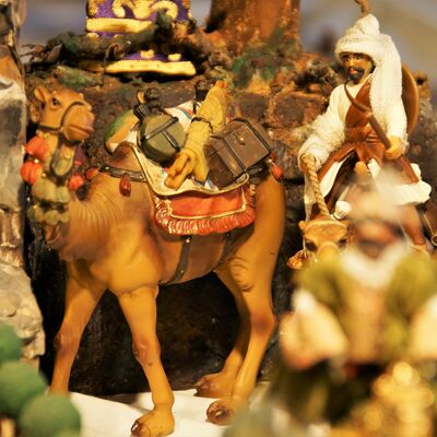 Bild vergrößern: Figuren aus der Krippe "Fünf Epiphanien als barockes Welttheater". Ein vollgepacktes Kamel steht vor einer menschlichen Figur in weißer Kleidung und Rute in der Hand.