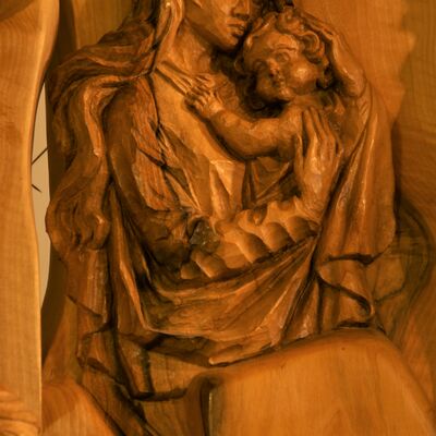 Bild vergrößern: In der MItte der aus Relief geschnitzten Krippe ist eine Frau mit langem Gewand und langen Haaren, welche ein Baby im  Arm hält. Die beiden Köpfe berühren sich.