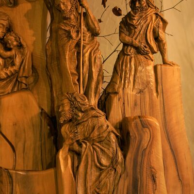 Bild vergrößern: Geschnitzte Relief Krippe. Drei in den Holzstamm geschnitzte Personen. Im Hintergrund ein Hirte mit Hirtenstab im Vordergrund kniet ein Mann mit Bart vor Maria und dem Jesuskind. Die dritte Person, ebenfalls männlich stützt sich auf einen Baumstamm.