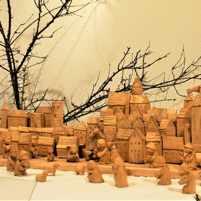 Bild vergrößern: Aufnahme einer Krippenlandschaft, welche aus Holz geschnitzt wurde. IM Vordergrund viele kleine aus Holz geschnitzte Figuren mit rundlicher Form. Im Hintergrund die Silhouette einer Stadt mit vielen verschiedenen Häusern. Großen, Kleinen, Hohen, Tiefen.