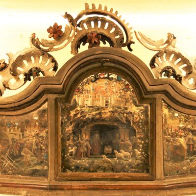 Bild vergrößern: "Rokoko Kastenkrippe" ist eine Krippe aus dem Jahr 1750. Die Front des "Kastens" ist in einer dreieckigen From aus Glas. Die oben mit vielen verschnörkelten Details verziert ist. Innen sieht man eine große Burg, sowie einen Wald mit verschiedenen Figuren (Menschen und Tieren)