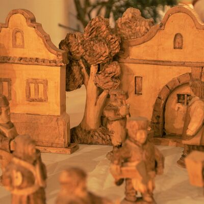 Bild vergrößern: Geschnitzte Holzkrippe. Das Holz weist Strukturen auf und ist unterschiedlich hell. Im Hintergrund sind zwei Häuser zu sehen. An ein Haus schmiegt sich ein Baum dich an. IM Vordgerund sind viele Figuren zu sehen. Jede Figur hat eine andere Aufgabe. Sie haben Mützen auf, tragen Körbe oder Holz in den Händen.
