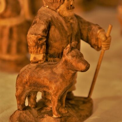 Bild vergrößern: Nahaufnahme einer geschnitzten Holzfigur und eines geschnitzten Tieres. Einem Hund. Der Besitzer des Hundes hat einen Stab in der Hand, hat einen Bart und eine Mütze auf.