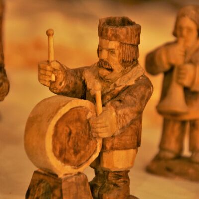 Bild vergrößern: Zu sehen ist eine aus Holz geschnitzte Figur mit Wintermantel und Mütze. Männlich und Schnauzbart. Vor der Fogur ist eine Trommel. Figur trommelt mit 2 Trommelstäben links und rechts auf die große Trommel.