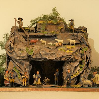 Bild vergrößern: Gesamtdarstellung der Krippe aus Distanz. Eine detaillreiche Krippendarstellung. Ein Berg mit einer Höhle. Über der Höhle auf dem Berg stehen Schaafe und Ziegen sowie zwei männliche Hirten.  In der Höhle ist eine weibliche Person und eine männliche Person dargestellt. Die männliche Person hat einen Stab in der Hand. In der Mitte der beiden Personen steht eine Krippe mit einem Baby darinnen.Rechts und Links neben dem Höhleneingang sind weitere Figuren abgebildet, welche alle Gaben in den Händen halten und demütig an die Höhle herantreten.