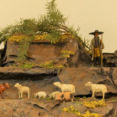 Bild vergrößern: Detailaufnahme des Geschehens auf dem Berg. Mehrere Schaafe grasen verteilt auf dem Berg. Im Hintergrund ein Hirte mit Hirtenstab und Hut und langem Mantel.