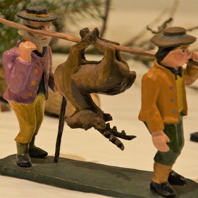Bild vergrößern: Die Krippenfiguren sind aus einer speziellen Modelliermasse hergestellt. Zwei Figuren mit Hut, klein, tragen ein an einem Stock aufgehngtes Totes Tier. Ein Reh.