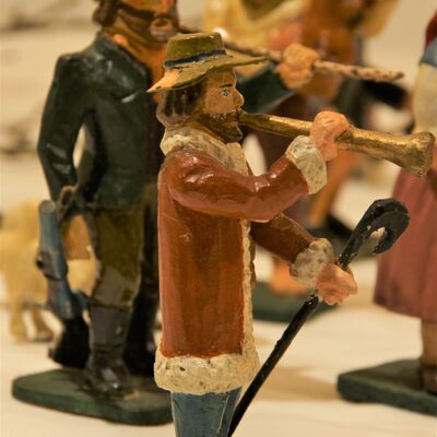 Bild vergrößern: Eine Figur mit einem Hut und in einem langen Wintermantel gekleidet. In einer Hand einen gebogenen Hirtenstock. Die Figur spielt Trompete. Im Hintergrund viele Figuren, unscharf.
