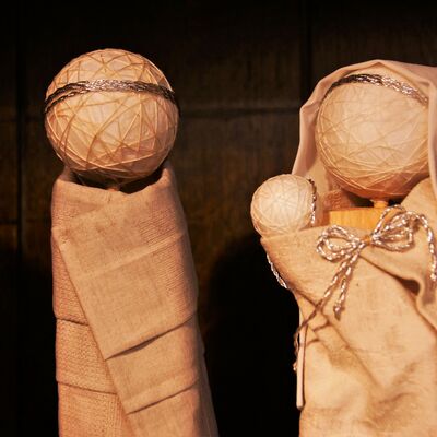 Bild vergrößern: Zwei Figuren. Sehr einfach gehalten. Der körper besteht aus einem kleinen Holzklotz. Auf Diesen ist ein Ball mit strukturen als Kopf gesetzt. Die Figur ist in einem Gewand eingekleidet und trägt auf dem Kopf einen Art Krone. Neben der Figur steht eine Weitere Figur, die zusätzlich ein Kopftuch trägt und eine kleine Klötzchenfigur, ein Baby, in den Armen hält.