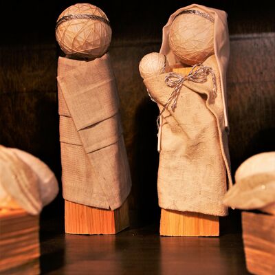 Bild vergrößern: Zwei Figuren. Sehr einfach gehalten. Der körper besteht aus einem kleinen Holzklotz. Auf Diesen ist ein Ball mit strukturen als Kopf gesetzt. Die Figur ist in einem Gewand eingekleidet und trägt auf dem Kopf einen Art Krone. Neben der Figur steht eine Weitere Figur, die zusätzlich ein Kopftuch trägt und eine kleine Klötzchenfigur, ein Baby, in den Armen hält. IM Vordergrund sind zwei Schaafe zu sehen.