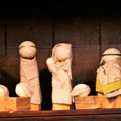 Bild vergrößern: Gesamt-Abbildung der Krippe. Vier Figuren sind nebeneinander aufgestellt. Die körper aus Holzklotzen die Köpfe als runder Ball mit Krone. Zwei Figuren tragen ein Kopftuch. Eine Figur trägt ein Baby im Arm. Im Vordergrund sind Schaafe, ebenso mit Holzklotz Körper.