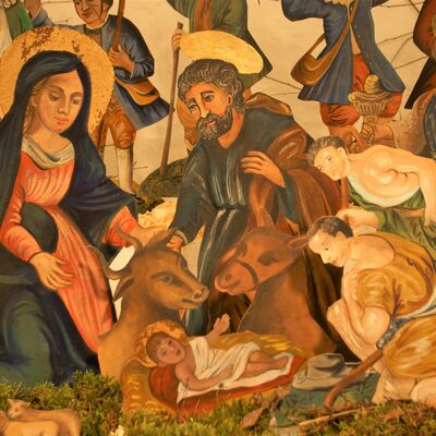 Bild vergrößern: Detailaufnahme mit den Figuren Maria, Josef und Jesuskind. Eine weibliche Person kniet zusammen mit einer älteren, männlichen Person, mit Bart hinter einer Krippe. Beide haben einen Heiligenschein über dem Kopf. In der Krippe liegt ein im Gewand gekleidetets Baby. Zwei Tiere, ein Esel und ein Ochse sind abgebildet. Aus dem rechten Bildrand lehnen sich zwei Männer in das Bild und bewundern das Baby.