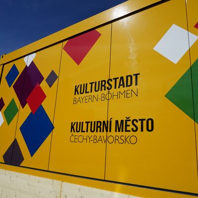 Bild vergrößern: Gelber Container mit den vielen verschiedenfarbigen Vierecken, die das Logo des Kulturstadtjahres zieren. Der Container ist teils aus Metall, teils aus Glas und begehbar.
