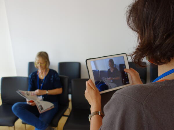 Bild vergrößern: Eine Frau mit einem Prospekt sitzt auf einem Stuhl. Eine weitere Frau filmt sie mit dem Tablet.