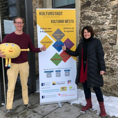 Bild vergrößern: Veronika Hofinger und David Bender stehen neben dem bunten Kulturstadt RollUp mit dem Maskötchen "der Knödel" da.