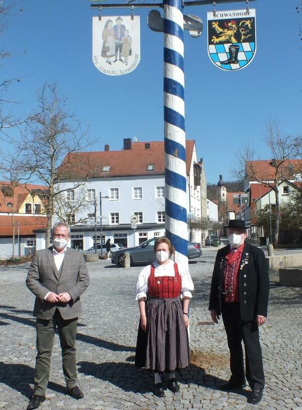 Bild vergrößern: Drei Personen vor einem Maibaum auf einem großen Platz.