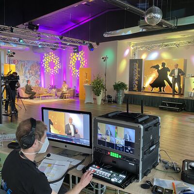 Foto von der Technik, die hinter dem Live-Stream steckt.
2 Bildschirme, viele Tasten und eine Person. Im Hintergrund sieht man die dekorierten Räume der Tanzschule Theuerl.