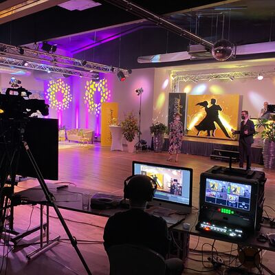 Eine Fotoaufnahme hinter den Kulissen.
Man sieht 2 Bildschirme, eine Person mit Kopfhörern, eine Filmkamera. Alles fokussiert auf die festlich dekorierten Räume der Tanzschule Theuerl.