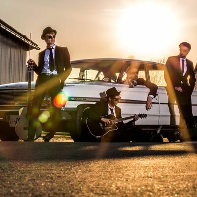 Bild vergrößern: Vier junge Männer posieren im Anzug bei Sonnenuntergang bei einem alten Auto. Zwei lehnen an dem Fahrzeug, einer sitzt vor dem Auto und der letzte innen drin.