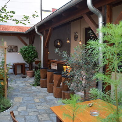 Bild vergrößern: Im Innenhof der Stube Edenharter befindet sich eine gemütliche Bar aus Holz und davor stehen vier Barhocker. Verschiedene Sträucher und Blumen, wie ein Olivenbäumchen, sind im gesamten Bereich verteilt.