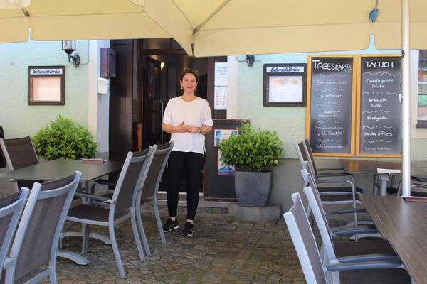 Im Außenbereich zwischen den Tischen und unterm Sonnenschirm steht die Inhaberin Frau Sojic. Im Hintergrund ist neben der Eingangstür eine dekorative Bepflanzung.
